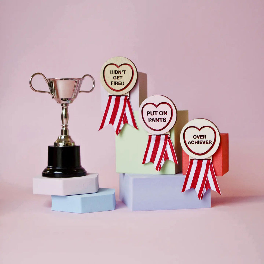 Candy & Kitsch candy heart merit badge brooch sits in a kitsch pink background with a trophy in the variation ’over achiever'