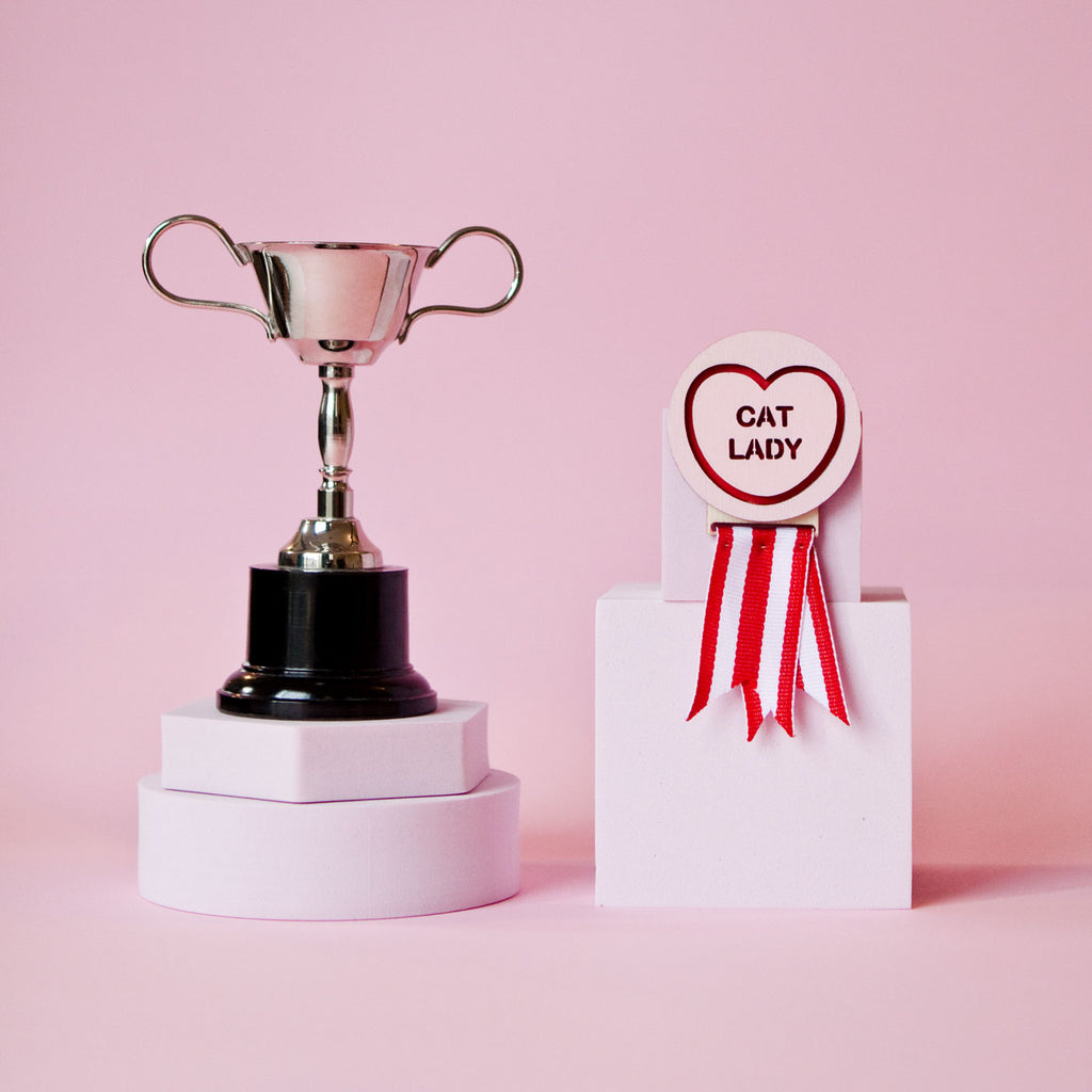 Candy & Kitsch candy heart merit badge brooch sits in a kitsch pink background with a trophy in the variation ’cat lady'