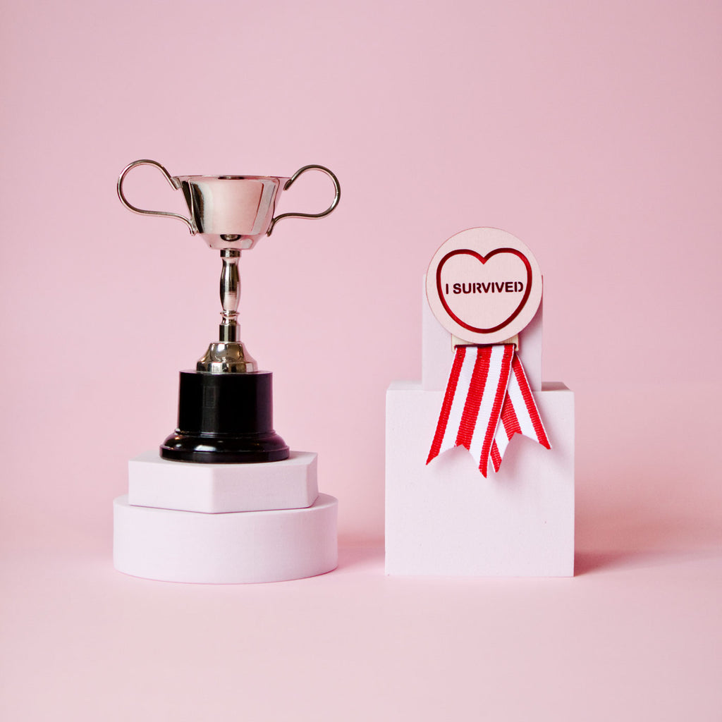 Candy & Kitsch candy heart merit badge brooch sits in a kitsch pink background with a trophy in the variation ’i survived'