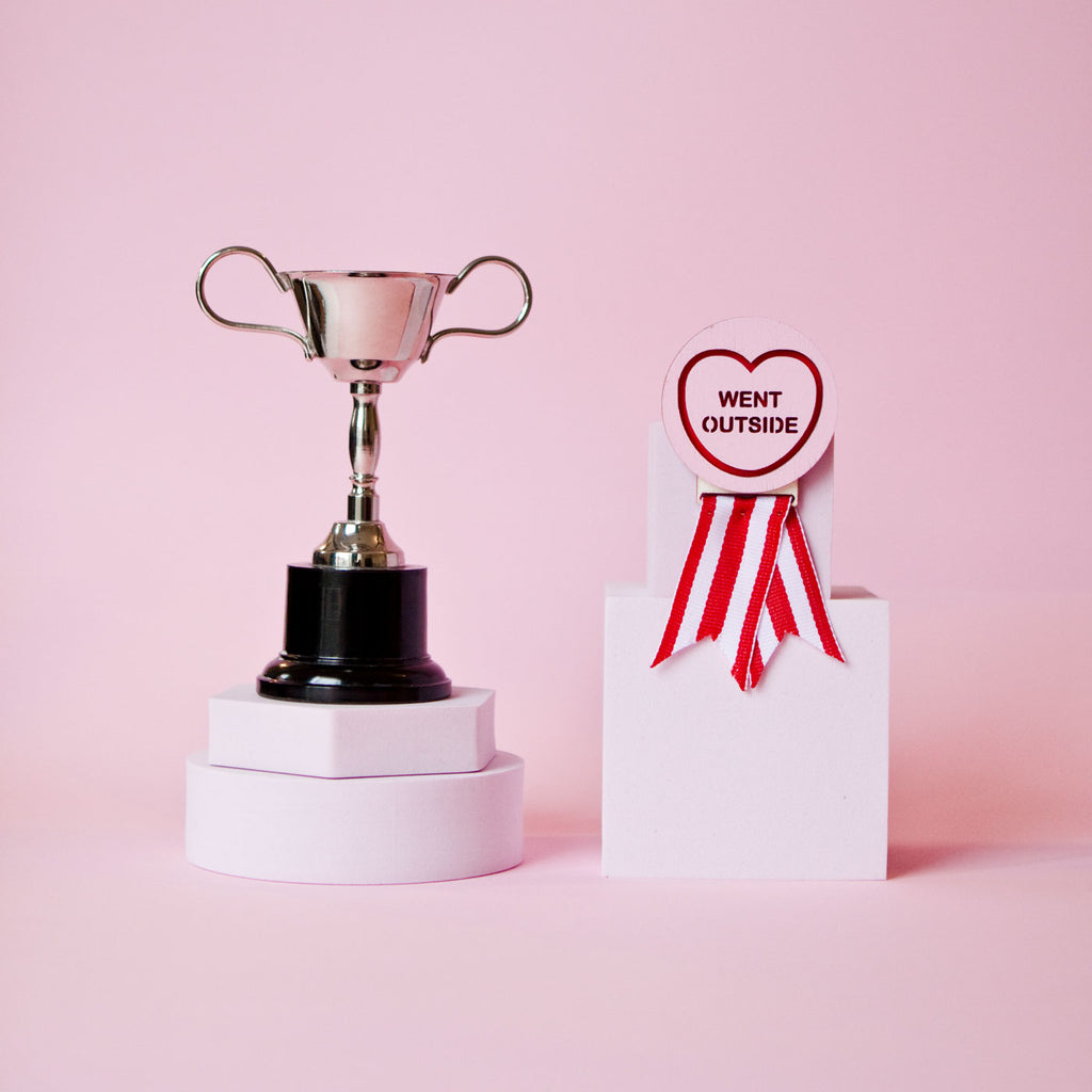 Candy & Kitsch candy heart merit badge brooch sits in a kitsch pink background with a trophy in the variation ’went outside'